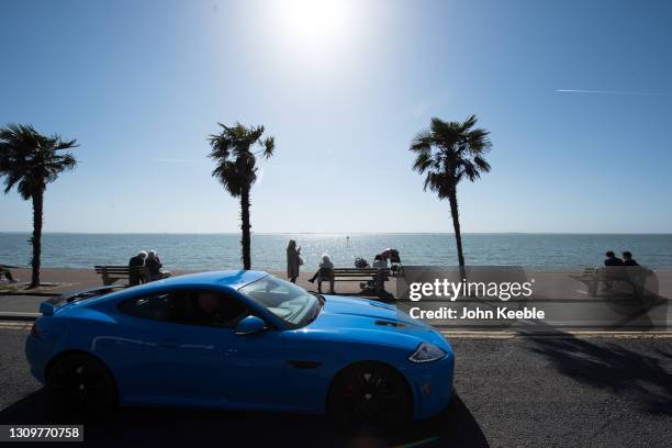 People sit on benches to enjoy the warm weather as a Jaguar XKR-S sports car passes by on the promenade beside the beach in the warm weather on March...