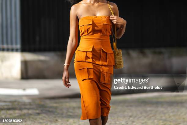 Magalie Kab wears long earrings, an off-shoulder orange dress with six attached pockets, a yellow grid bag, on March 28, 2021 in Paris, France.