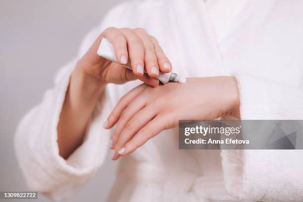 woman in white bathrobe with nude pink manicure holds tube with natural organic serum. she is squeezing cream on hand's skin to moisturize it. concept of home body care and healthy lifestyle. close-up front view - creme tube ス�トックフォトと画像