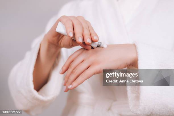 woman in white bathrobe with nude pink manicure holds tube with natural organic serum. she is squeezing cream on hand's skin to moisturize it. concept of home body care and healthy lifestyle. close-up front view - hand cream 個照片及圖片檔