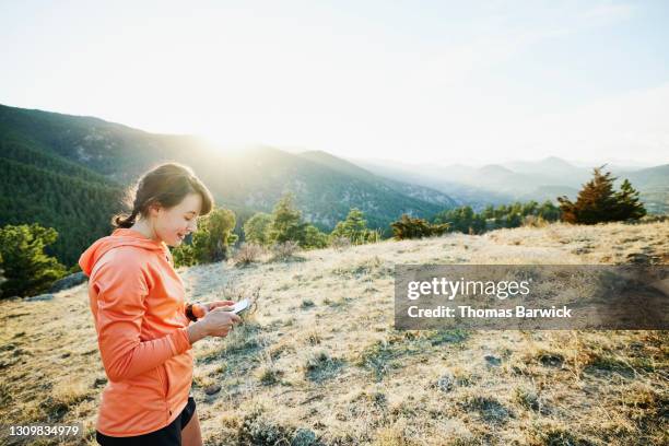 medium wide shot of smiling female athlete sending picture from smart phone after trail run in mountains - co op stock pictures, royalty-free photos & images