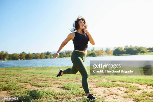 smiling young woman jogging near lake on sunny day - jogging stock-fotos und bilder