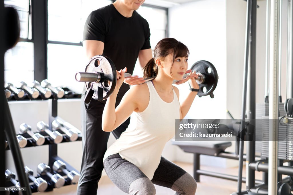 Asian woman doing barbell squats with the assistance of a trainer