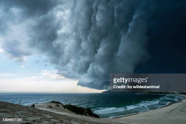 two people sitting on top of a sand dune watching a large storm front moving across one mile beach, forster, nsw, australia. - thunderstorm stock-fotos und bilder