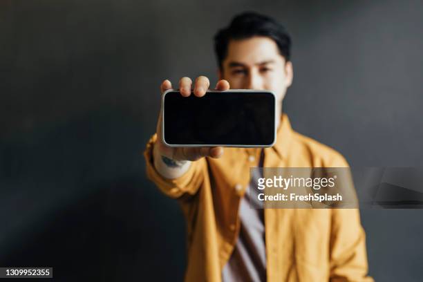 giovane bell'uomo che indossa una camicia gialla, mostrando lo schermo del suo smartphone alla fotocamera - mostrare foto e immagini stock