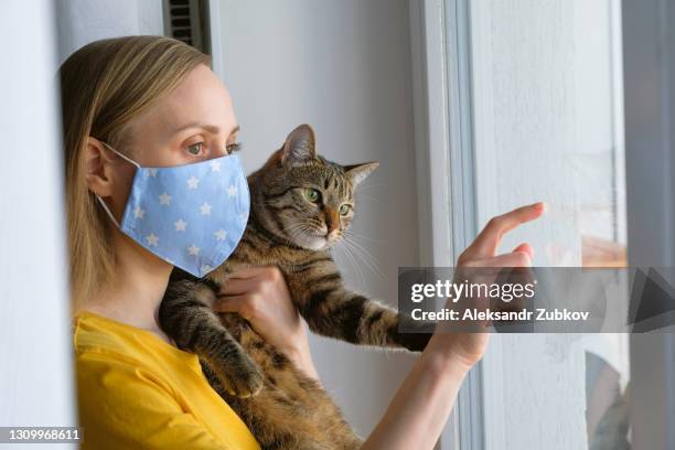 the woman put on a reusable protective mask for home isolation from coronavirus and covid-19. a girl holds a cat in her arms, indoors or at home by the window. preventing the spread of the virus and disease. waiting for the quarantine to complete. - cat face mask stock-fotos und bilder