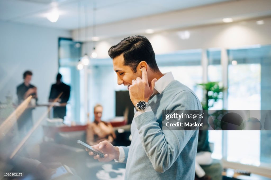 Happy businessman using smart phone while adjusting earphones in creative office