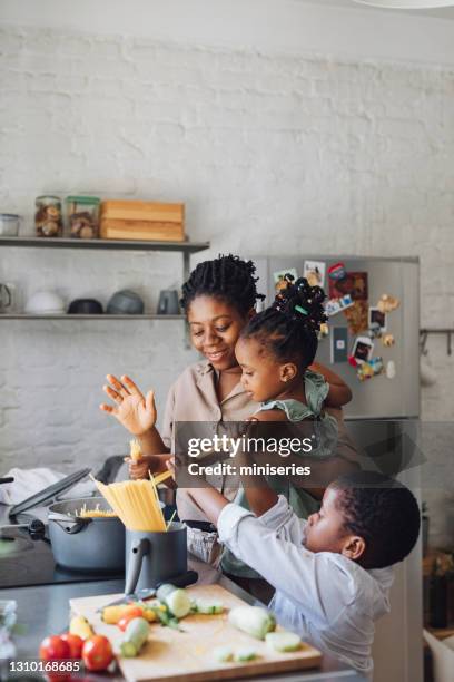 mutter, tochter und sohn bereiten spaghetti und gemüse für das mittagessen über einem schneidebrett - black mother and child cooking stock-fotos und bilder