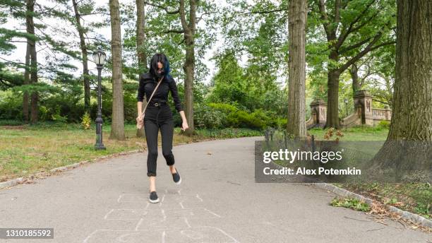 a teenage girl playing hopscotch in central park, manhattan, new york, deserted because of the covid-19 pandemic. she is wearing a protective mask but holds in just on the neck because nobody is around. - hopscotch stock pictures, royalty-free photos & images