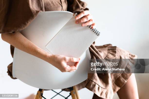 the girl sits on a white chair with wooden legs and holds a notebook in her hand. - brown dress stockfoto's en -beelden