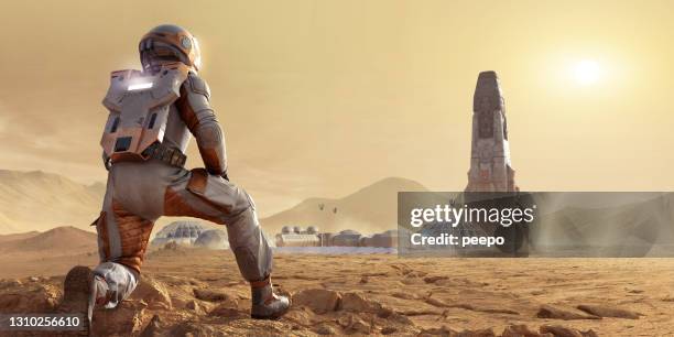 astronaut on mars kneeling and looking at base camp settlement and rocket in mars rocky environment - basislager stock-fotos und bilder