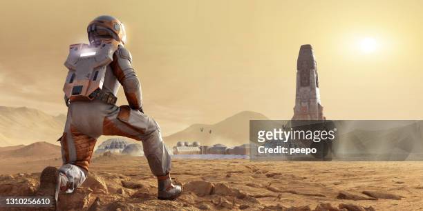 astronaut op mars knielend en kijkend naar basiskamp nederzetting en raket in mars rotsachtige omgeving - spaceship stockfoto's en -beelden