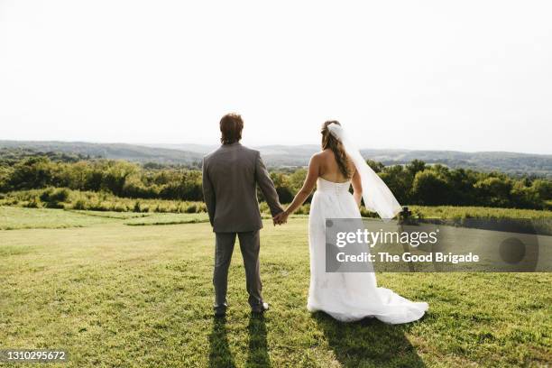 rear view of newlywed couple holding hands in grassy meadow - rural scene wedding stock pictures, royalty-free photos & images