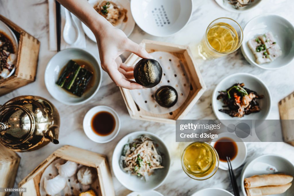 Flat lay of assorted traditional Chinese dim sum in bamboo steamer with a variety of appetitzers freshly served on table with a woman enjoying meal and holding a steamed bun in restaurant. Chinese cuisine and food culture. Yumcha. Eating out lifestyle