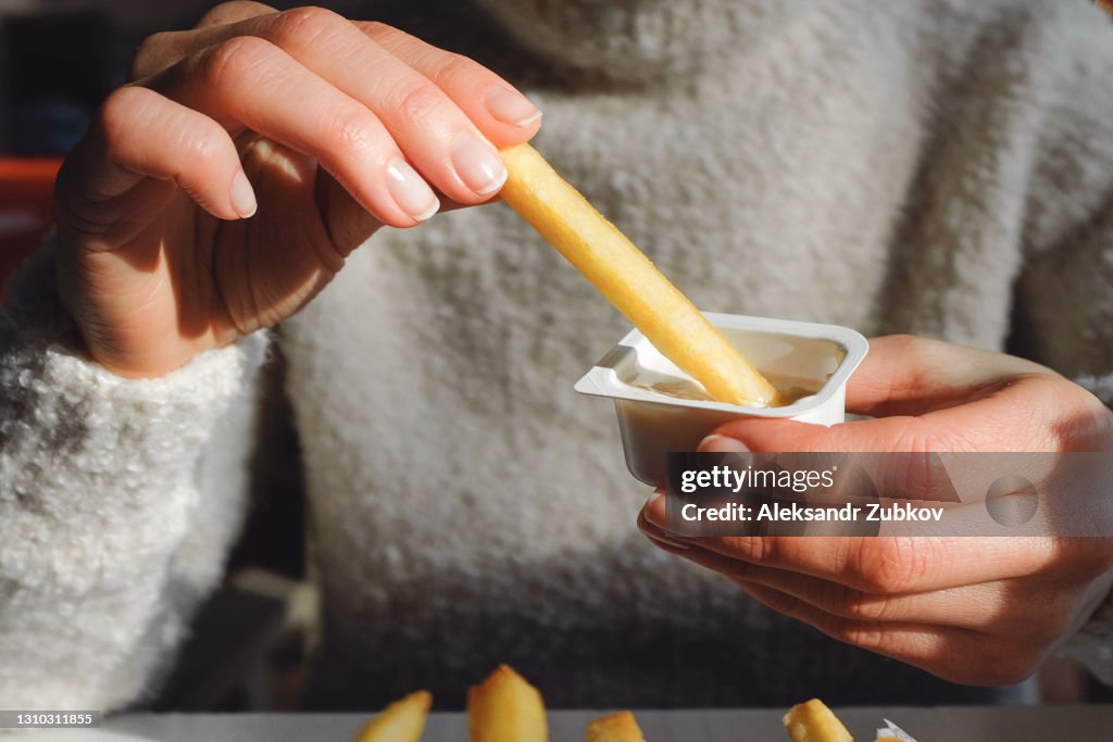 A woman holds French fries in her hand, eating them in a cafe or restaurant. A girl in a white knit sweater dips a slice of potato in garlic sauce. The concept of fast food, snacking, junk food.