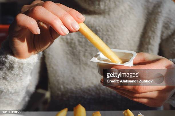 a woman holds french fries in her hand, eating them in a cafe or restaurant. a girl in a white knit sweater dips a slice of potato in garlic sauce. the concept of fast food, snacking, junk food. - mojar fotografías e imágenes de stock