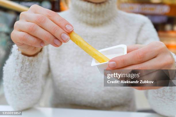 a woman holds french fries in her hand, eating them in a cafe or restaurant. a girl in a white knit sweater dips a slice of potato in garlic sauce. the concept of fast food, snacking, junk food. - girl eating french fries stock pictures, royalty-free photos & images