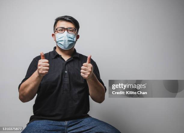 studio shot of an asian man wearing face mask sitting with both thumbs up - 手のしぐさ ストックフォトと画像