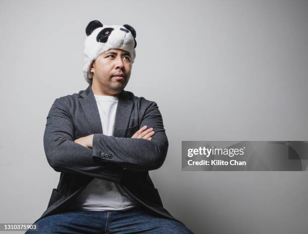 studio shot of an asian man wearing panda hat sitting with arms crossed - asian man studio shot stock pictures, royalty-free photos & images