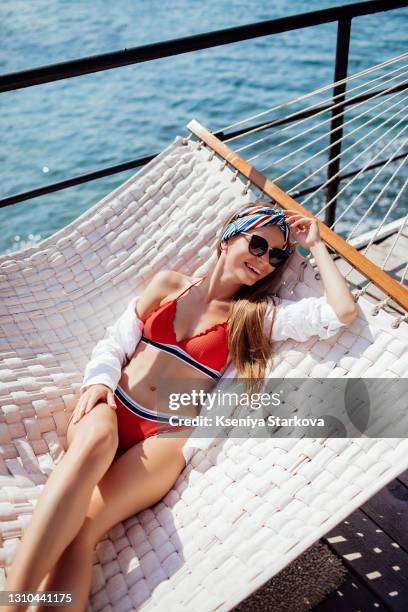 young woman with long blond hair lies in a red swimsuit on a white hammock on the seashore - red swimwear stock pictures, royalty-free photos & images