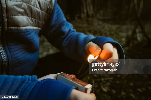 a mischievous child in the dark outdoors holds a burning match and a matchbox in his hands. danger of fire for children. risk of burns and forest fires - arson foto e immagini stock