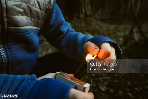 a mischievous child in the dark outdoors holds a burning match and a matchbox in his hands. danger of fire for children. risk of burns and forest fires - arson stock pictures, royalty-free photos & images