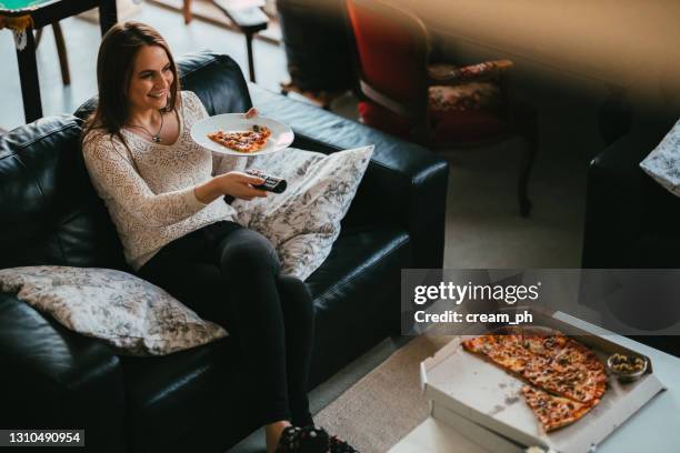 junge lässig gekleidete frau, die pizza vor einem fernseher isst - zu hause bleiben redewendung stock-fotos und bilder