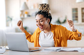 Overjoyed excited african american girl with dreadlocks, freelancer, manager working remotely at home using laptop, looks at screen with surprise, smiling face, gesturing with hands, got a dream job