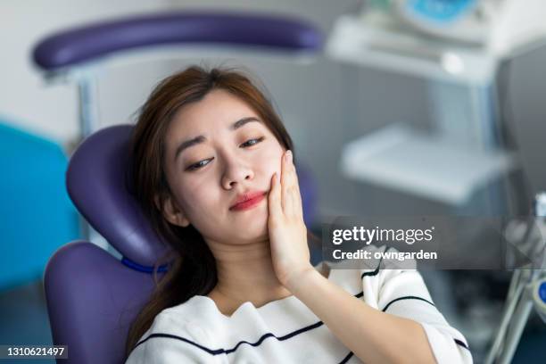 young woman holding cheek in chair at dentist, having toothache. - toothache stock pictures, royalty-free photos & images
