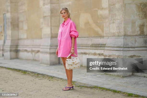 Xenia Adonts wears golden earrings, a neon pink long oversized shirt / dress from Valentino, a white Valentino studded bag, a golden watch, a golden...