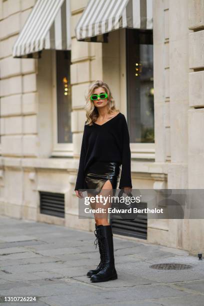 Xenia Adonts wears neon green sunglasses from Chiara Ferragni, a black wool pullover with v-neck from Bazilika, a black short mini shiny leather slit...