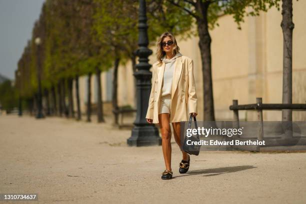 Xenia Adonts wears sunglasses, a beige oversized blazer jacket from Enveloppe, a white wool hoodie sweater and shorts from Uzwei, a black leather...