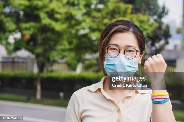 portrait of asian woman holding a fist with hand wearing "covid-19 vaccinated" rubber wristbands - rubber bracelet stock pictures, royalty-free photos & images