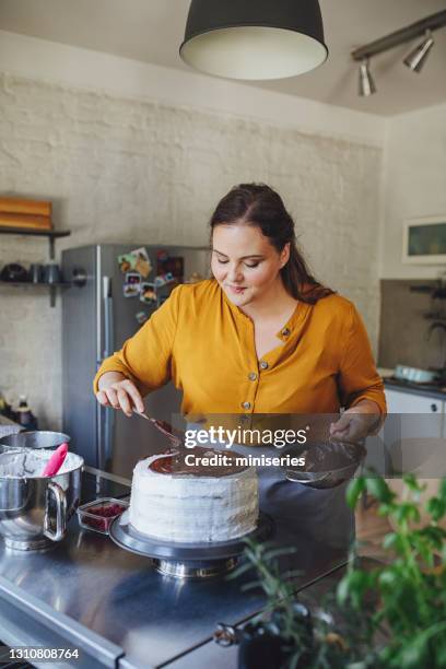 cheerful young caucasian woman spreading chocolate over the cake - chocolate cake stock pictures, royalty-free photos & images