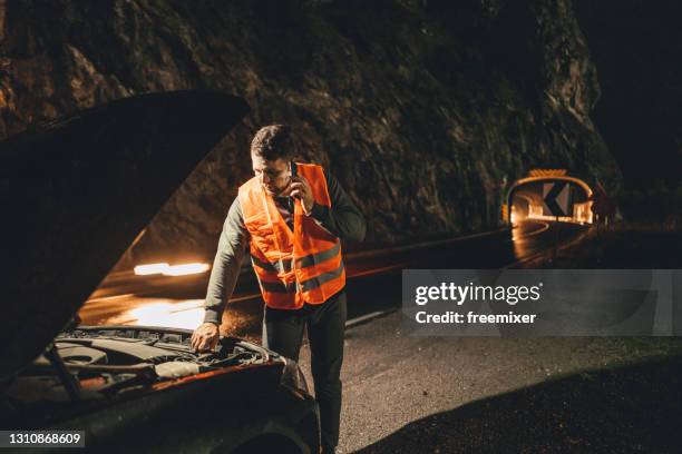 male driver in reflective waistcoat looking in car hood on roadside while talking with roadside service on phone - assistência na estrada imagens e fotografias de stock