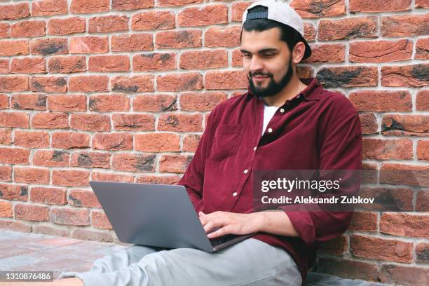 an east indian american college student is sitting outside, working on a computer or laptop. happy hindi man chatting with friends or loved ones via video link, smiling, against the background of a brick wall of a building. - netbook stock-fotos und bilder