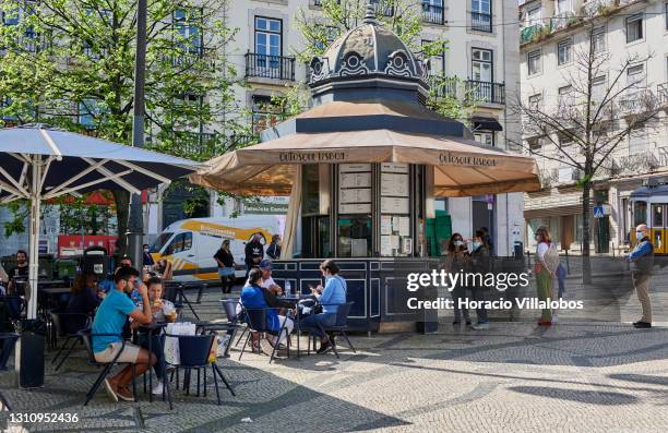 Mask-clad patrons queue for food and drinks while others sit at tables at Quiosque Lisboa in Praça Luís de Camões on the first day of shops reopening...