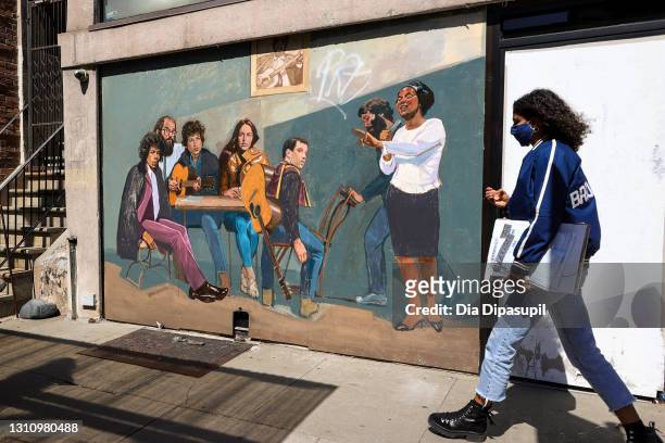 Woman wearing a protective mask walks past a mural titled "Spirit of Greenwich Village" by Amer Olson featuring Jimi Hendrix, Allen Ginsberg, Bob...