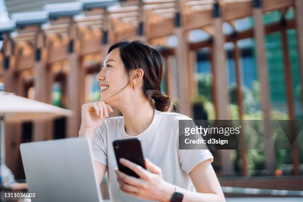 confident young asian freelance businesswoman looking away with smile while using smartphone and working on laptop on the go outdoors in urban park in the city. remote working concept with flexible lifestyle - woman using smartphone with laptop stock-fotos und bilder