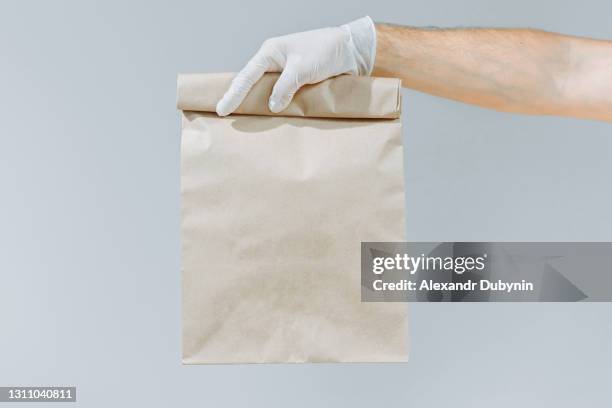 hand of a man in a glove holding a brown bag or bag for taking out isolated on a gray background. packaging layout template. delivery service concept. copy space. courier - sachet en papier photos et images de collection