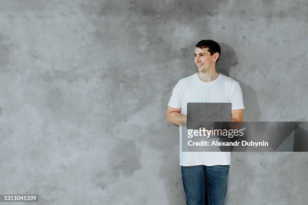 young man standing and holding laptop computer looking to side on gray background in studio - pleased face laptop stock pictures, royalty-free photos & images