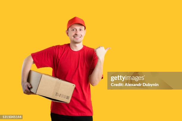 online home delivery service. a courier man in a red t-shirt and a cap holds a cardboard box in his hands and shows his finger in copy space isolated on a yellow background in the studio. - white shirt template stock pictures, royalty-free photos & images