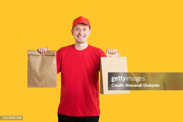 food delivery service from an online store. courier in red uniform with craft bags gives fast food order on yellow isolated background. copy space - take away food courier stock pictures, royalty-free photos & images