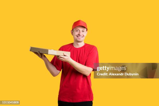 pizza delivery service. happy courier in red uniform holds pizza in hands on yellow isolated background in studio. copy space - pizza delivery stock-fotos und bilder