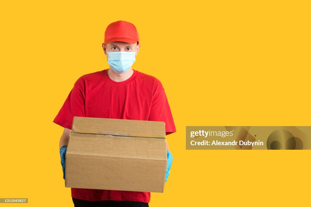Food delivery service. delivery employee in a red cap and t-shirt, in a mask and gloves, holds a corona box on a yellow isolated background studio.