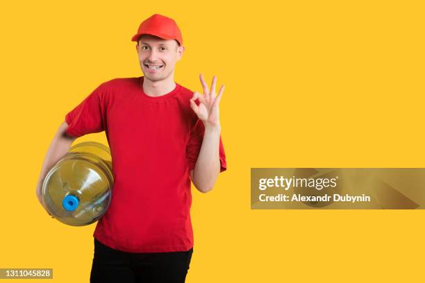 water delivery. happy courier in red uniform on yellow background with bottle of water shows ok sign isolate. copy space - red polo stock pictures, royalty-free photos & images