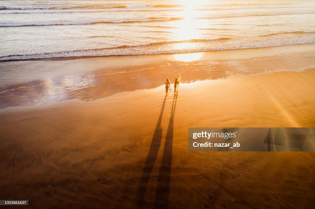 Two women carrying surfboard on beach at sunset time.