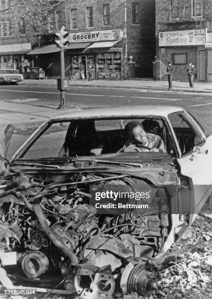 Young African American boy playing at the wheel of an abandoned car, which has had its front section stripped, possibly for spare parts, in the...