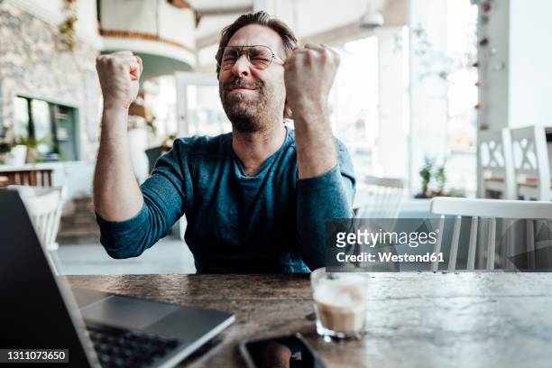 happy male entrepreneur doing fist by laptop in coffee shop - vincere foto e immagini stock