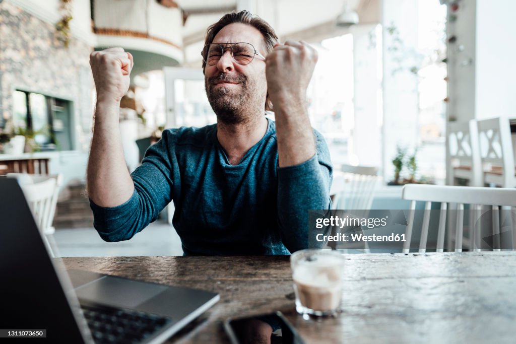 Happy male entrepreneur doing fist by laptop in coffee shop